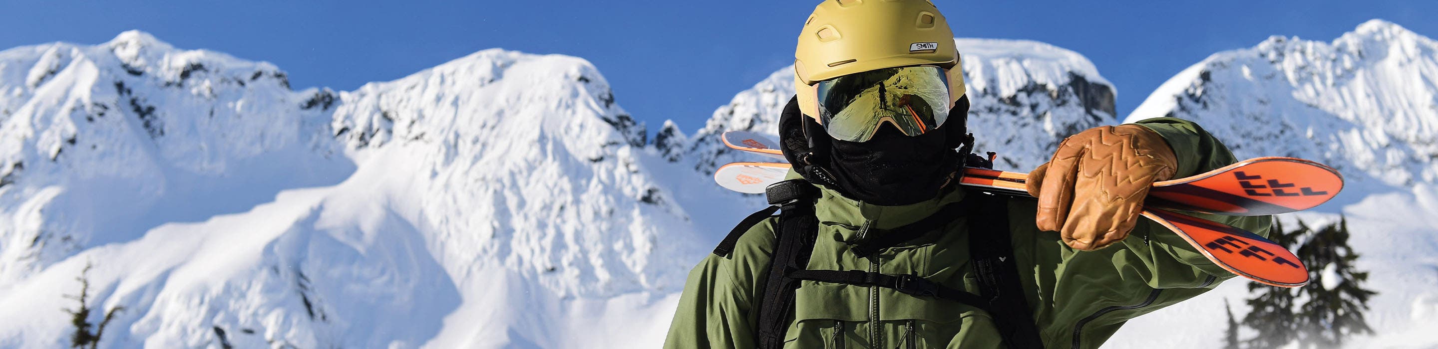 A man wearing OTG goggles poses for the camera with his skis over his shoulder with snowy mountains in the background.