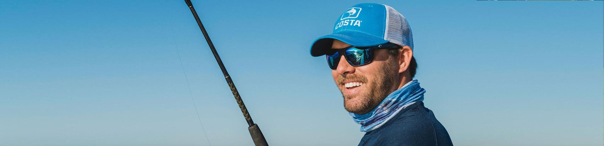An angler smiles as he looks into the sun in blue polarized fishing sunglasses, a blue neck gaiter, and blue long-sleeved t-shirt with his fishing rod in view.