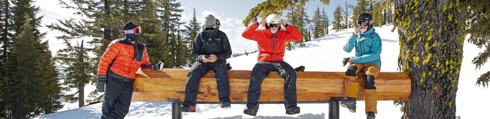 A few guys wearing prescription ski goggles for men take a break and sit on a log on a bluebird day in the snow.