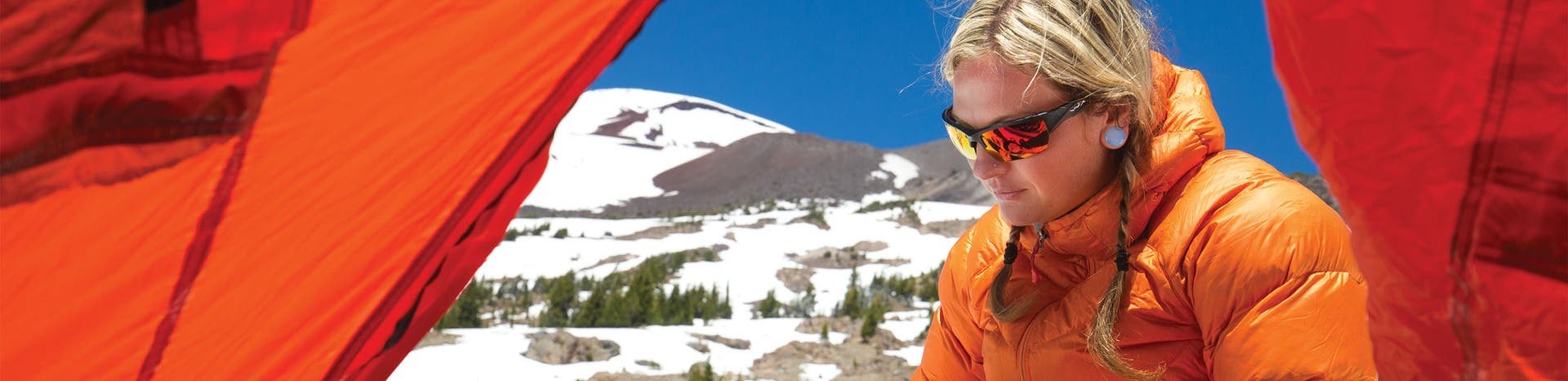 A young blonde woman in wrap around prescription sunglasses and an orange parka sits against snowy mountains and blue skies.